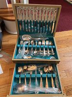 Full view of open original case containing knives in clipped slots on upper chamber and forks, spoons, and serving pieces in lower section, all with ornate silverplate handles and black blades on knives