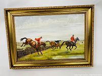 Framed oil painting of a fox hunting scene showing riders jumping a hedge with hunting dogs running alongside, signed by Unity Bush.