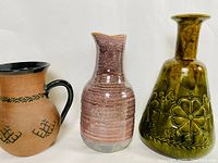Image showing three pottery pieces: a brown pitcher with black handle and geometric decoration, a pink and gray glazed vase, and a green glazed vase with embossed floral motifs.