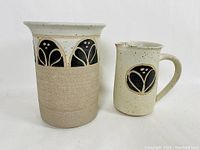 Front view of the wine cooler and pitcher side by side, showing size difference and decorative black motifs on neutral pottery clay.