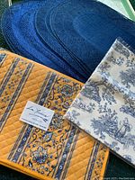 Photo showing four yellow quilted floral placemats, one packaged toile table runner with blue and cream toile design, and several navy woven oval placemats.