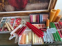 Overview of various boxed and loose new taper candles in red, white, orange, beige colors and multicolor boxed sets. Also shows green candles and clear glass candle holders.