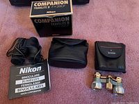 Photo showing Nikon Travelite III 7x20 CF binoculars, their box, a Nikon branded soft case, a MultiTech Survivor protected case, and Nikon booklet/manual laid out on a fabric surface.