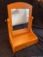 Small wooden dressing tilt mirror with visible drawer front and metal knob, showing mirror surface reflecting room elements.