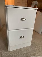 Front and side views of the white vertical file storage chest showing two drawers with silver cup handles and smooth white finish.
