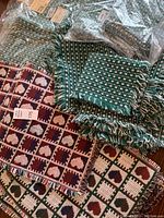 Group of table linens stacked including dark red placemats with heart patterns, dark green placemats with white weaving and fringes, and packaged green napkins.