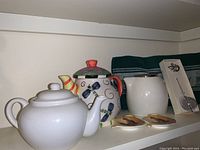 Two ceramic teapots (one white, one colorful), tea towel, tea infuser in box, and two ceramic teabag holders on shelf