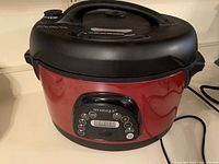 Front and side view of the red oval electric pressure cooker with black lid and handles on countertop.