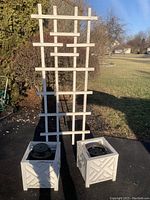Front view of a white wooden garden trellis behind two square white wooden planter pots with lattice pattern and black plastic liners. The items show peeling paint and outdoor wear.
