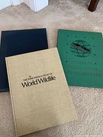 Three vintage hardcover atlases placed on carpet: Rand McNally Atlas of World Wildlife, New York Times Atlas of the World, and Colliers World Atlas and Gazetteer.
