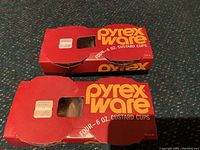 Two original red and yellow Pyrex Ware branded boxes each holding four custard cups, labeled 'FOUR - 6 OZ. CUSTARD CUPS'. Boxes show normal vintage wear.