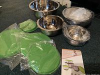 Four stainless steel mixing bowls of different sizes, each with silicone coated non-slip bottoms and separate green silicone lids, with packaging and instructions visible.