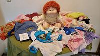 Full lot of Cabbage Patch Kid doll sitting on table surrounded by vintage and homemade clothes of various colors and types