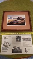 Framed print of a 1932 Ford car with barn and mountainous background as well as artist information sheet laid out below.