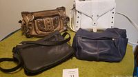 Photo showing set of four handbags on green surface, including Montana West white rhinestoned purse, tan and brown purse with studs and rings, dark gray leather bag, and navy blue purse.