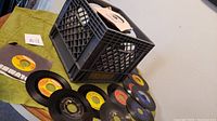 Photo showing a black milk crate with many 45 RPM vinyl records. Some records laid out on a table showing various colored labels.