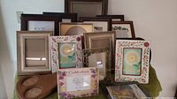 Various picture frames arranged on a green cloth background, showing different styles including floral designs, wooden, and gold ornate frames.
