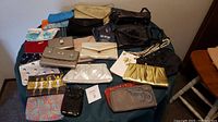 Photo showing 19 small bags, clutches, and purses arranged on a table with a chair beside it.