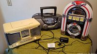 Three radios and CD players: cream Philips radio/CD player, brown Philips radio/CD cassette player, and Crosley jukebox lookalike radio/CD player on table