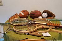 Multiple sports items including four wooden tennis rackets, two badminton rackets, two baseball gloves, and several sports balls including footballs, baseballs, and softballs spread on a table.