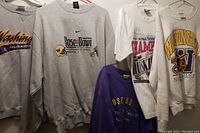 Five vintage UW Huskies shirts and sweatshirts hanging on hangers showing front designs including 1991 National Champs and 2001 Rose Bowl themes.