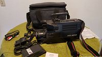 Wide shot showing the camcorder with lens extended, carrying case behind it, battery pack to the side, and connecting cables laid out on surface.