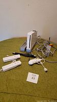 Full view of Nintendo Wii console missing top panel with two gray stands, two Wii remotes, one nunchuck, controller sensor bar, and cables on a green cloth background