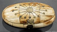Oval-shaped hand carved bone trinket box with circular metal inlay decoration on top, hinged lid and clasp.