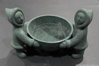 Front angle view showing the bowl held by two Inuit style figures with hooded parkas, in greenish-grey pottery material.