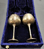 Top-down view of two tarnished silverplated wine goblets resting in a blue velvet-lined presentation box, showing exterior wear and dents.