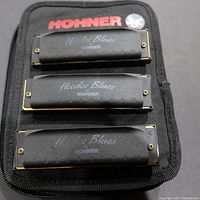 Top view showing three black Hohner HooDoo Blues harmonicas stacked on a black Hohner pouch with red logo.