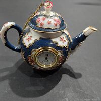Front view of the small blue and white porcelain teapot with pink floral accents, gold trim and a small clock embedded in the front.