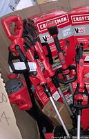 Photo of three red Craftsman V20 cordless lawn tools including string trimmers and blower vacuum near cardboard box and packaging.