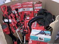 Wide view of lawn care tools standing in a box showing 2 string trimmers and 1 leaf blower/vacuum. Several Craftsman V20 Combo Kit branded boxes surround the tools.