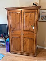 Front view of large wooden armoire with two paneled doors, black metal handles, and carved edges. Medium brown warm wood with knotty texture and visible scratches.