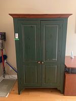 Front view of green painted wooden armoire with natural wood crown molding top, two doors with wooden knobs, standing on hardwood floor. Shows wear and scratches on painted surfaces.
