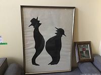 Photo showing both the large framed serigraph and smaller framed painting together on a wall, serigraph displays two abstract black figures.