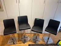 Four black vinyl bar stools arranged side by side showing full front and side views against a white closet background on a hardwood floor.