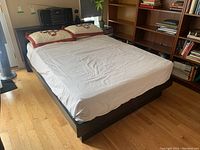 Painted wooden bed frame and headboard from a side angle showing the bed structure with white fitted sheet and pillows, in a room with wooden floors and nearby book shelves.