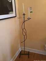 Photo of tall metal candlestick holder with two white taper candles, slender black metal frame with decorative looping design and rectangular base, situated in corner against yellow wall.