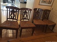 Photo showing four wooden chairs with decorative carved backs and rush seats arranged in a row on a hardwood floor.