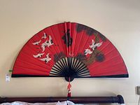 Front view of the large decorative wall fan mounted on a wall, showing the full red background, crane design, and tassel detail.