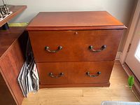 Front view of wooden filing cabinet showing two large drawers with two metal pull handles each, lock on top drawer, medium brown finish, standing on hardwood floor near window.