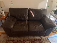 Front view of a brown leather love seat with two seats and cushioned armrests, showing some wear and creasing on the seats and arms.