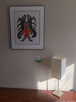 Framed painting on wall above wooden surface, depicting figure with black bird forms; table lamp with rectangular textured white fabric shade on metal base on surface beneath painting.