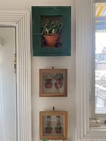 Photo showing three shadow box art pieces hung on a white wall by a window and door trim. Larger green frame contains an artificial plant in a terracotta pot. Two smaller wooden framed boxes contain images of multiple potted plants.