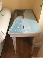 Side view of a wooden side table featuring a detailed hand-painted coastal scene on top, showing water, boats, and trees.