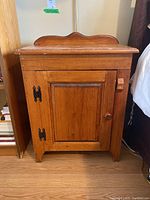 Front and side view of a wooden nightstand with a scalloped backboard, wooden door with wrought iron hinges and a knob on a hardwood floor.