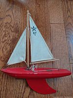 Side profile of red hull and collapsible white sails marked "Star SY 1" showing metal keel and rigging details on wooden floor.