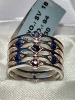 Five-band silver ring with seven blue sapphires displayed on white fabric, showing label indicating size and marking.